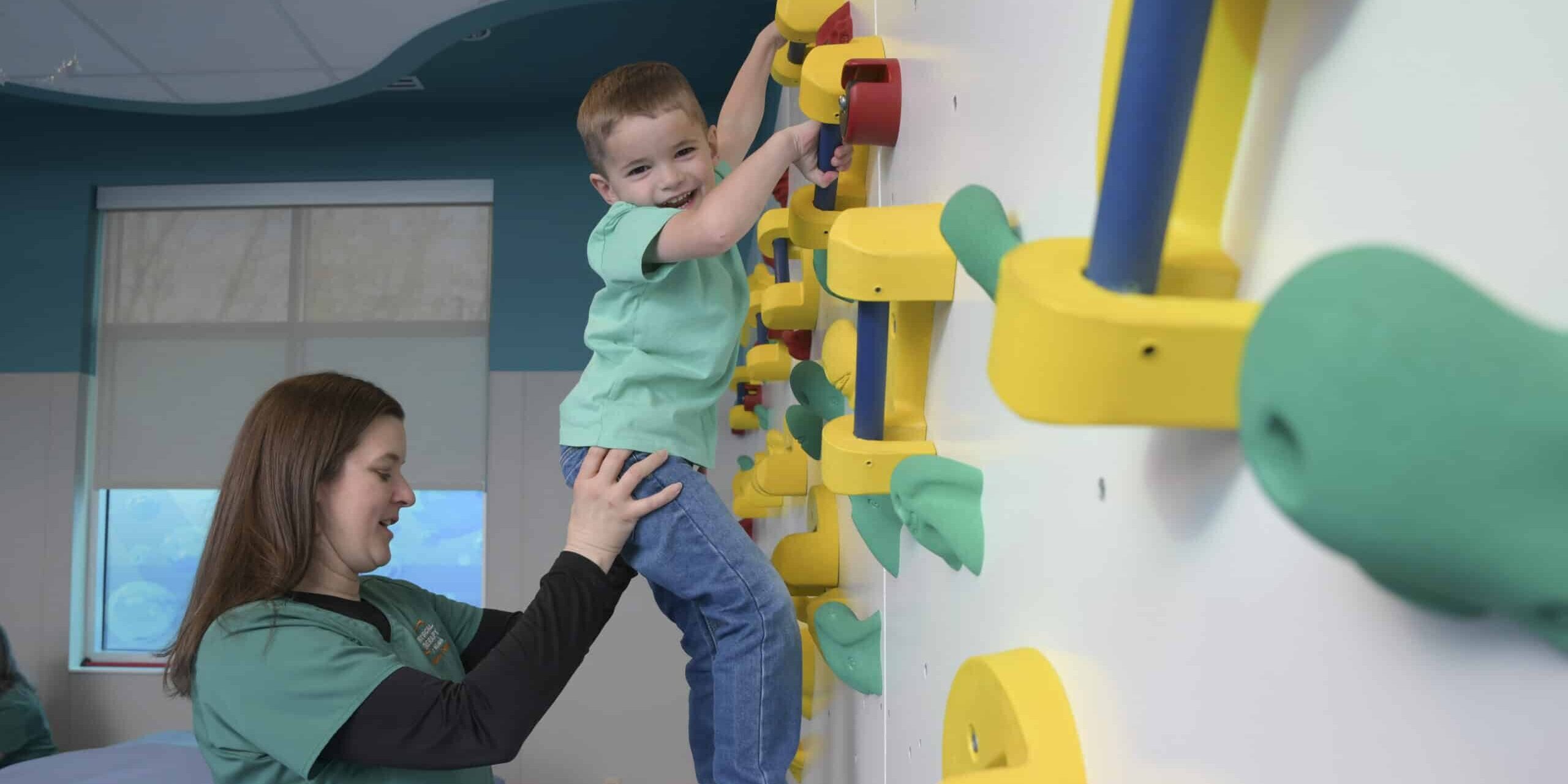 One of our expert pediatric physical therapists supports a pediatric patient as he climbs a child-safe rock wall, in an exercise to improve motor skills, hand-eye coordination, and mobility