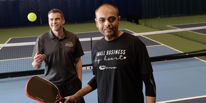 Mustafa Jaffer, a pickleball patient, stands in the foreground with a racket; behind him, Rick Grubb, expert physical therapist, tosses a pickleball ball